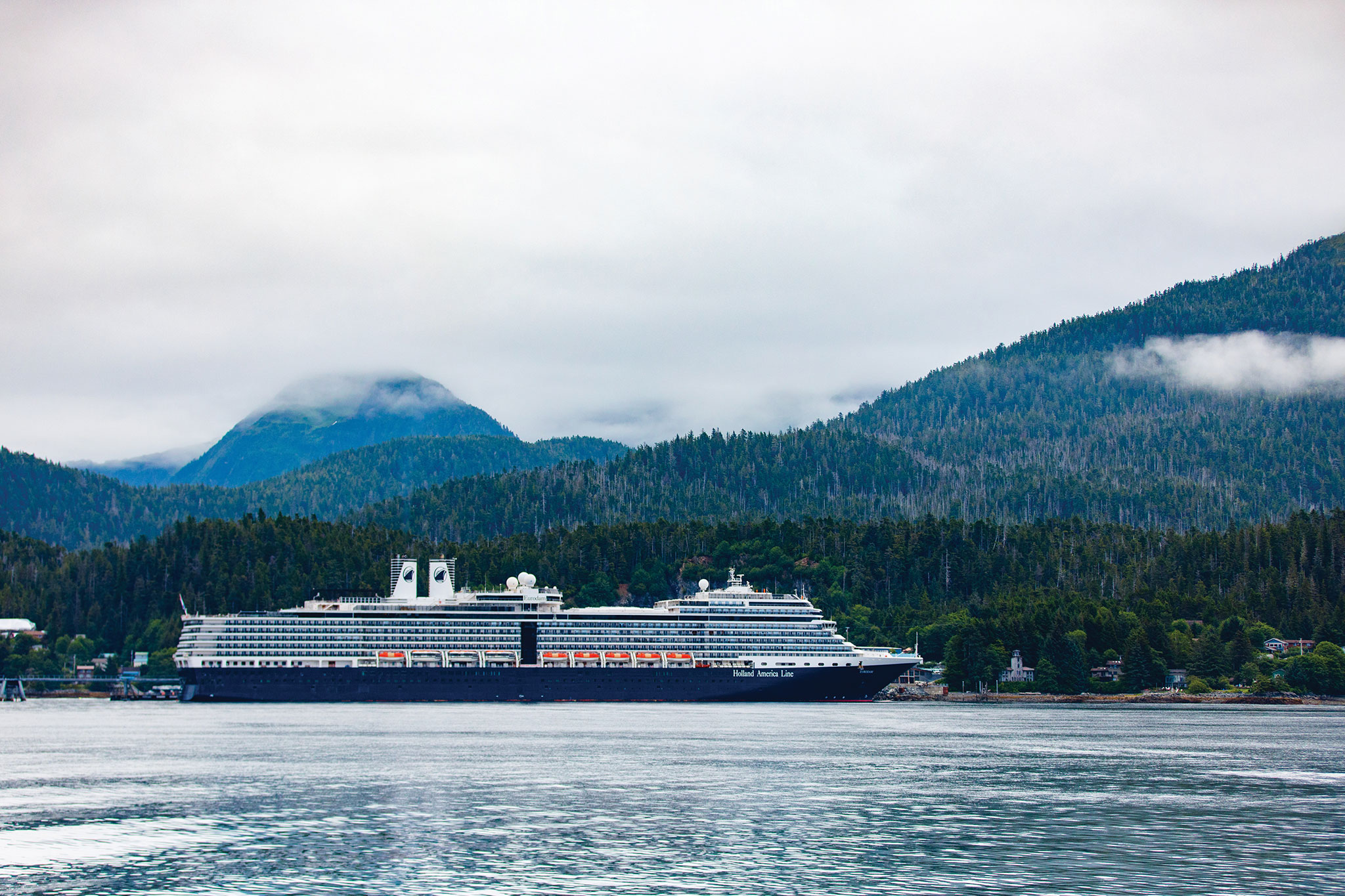 A cruise ship rests by a verdant coastline, surrounded by densely forested hills and misty mountains partly covered by clouds.