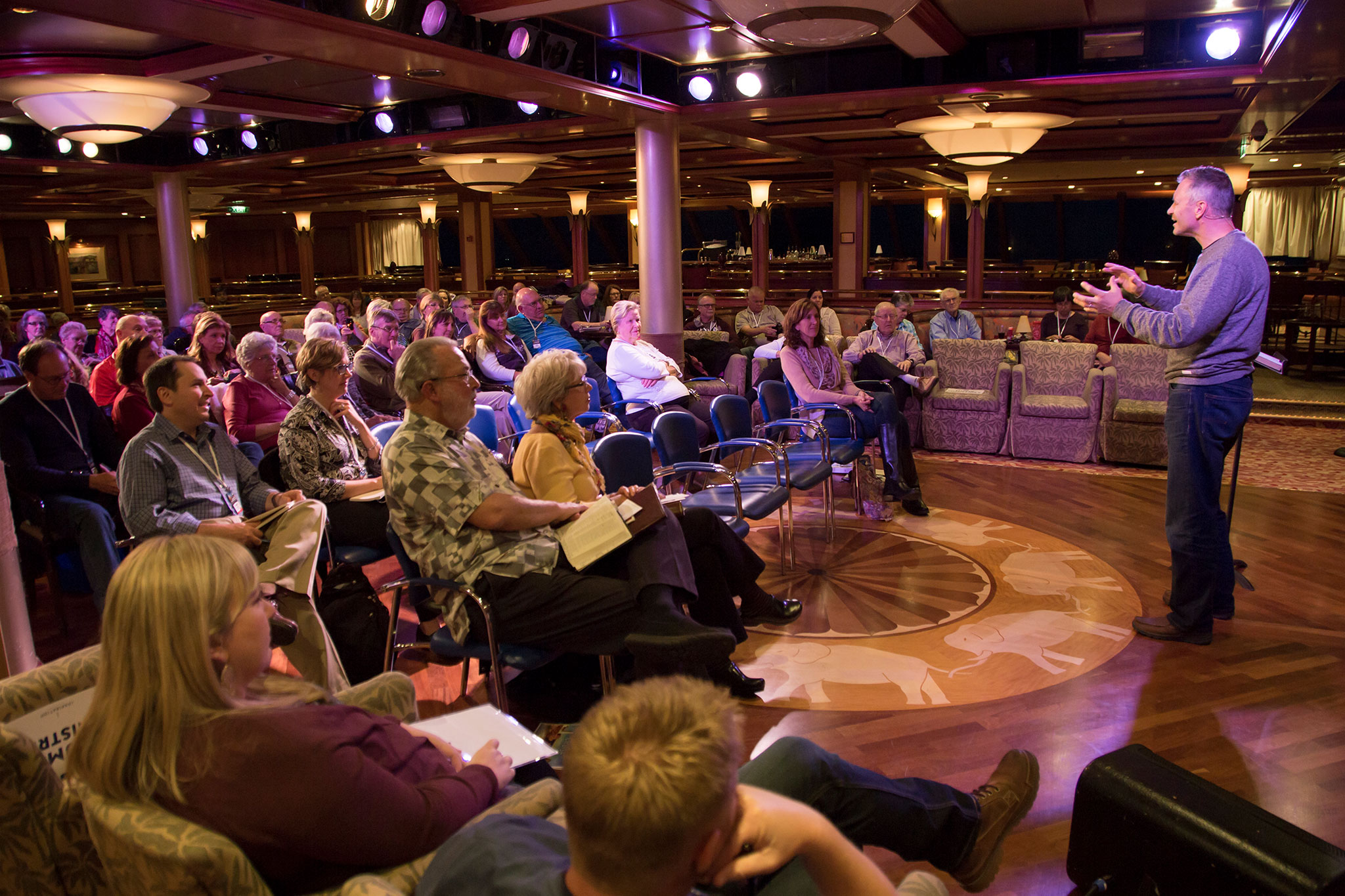 An engaging lecture in progress on a cruise ship, showing an audience of attentive guests listening to a speaker who is gesturing with his hands in the ship's elegantly designed lounge area.