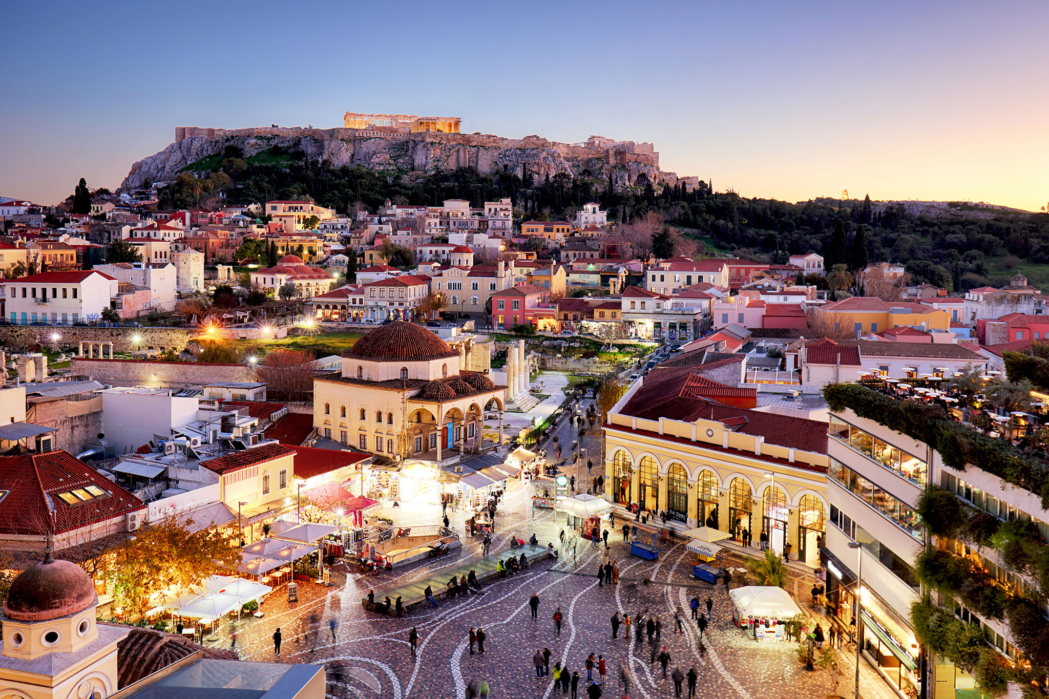 This is an aerial view of Monastiraki Square in Athens, Greece, at dusk. It shows bustling crowds, open-air markets, and the illuminated Acropolis on the hilltop in the background.