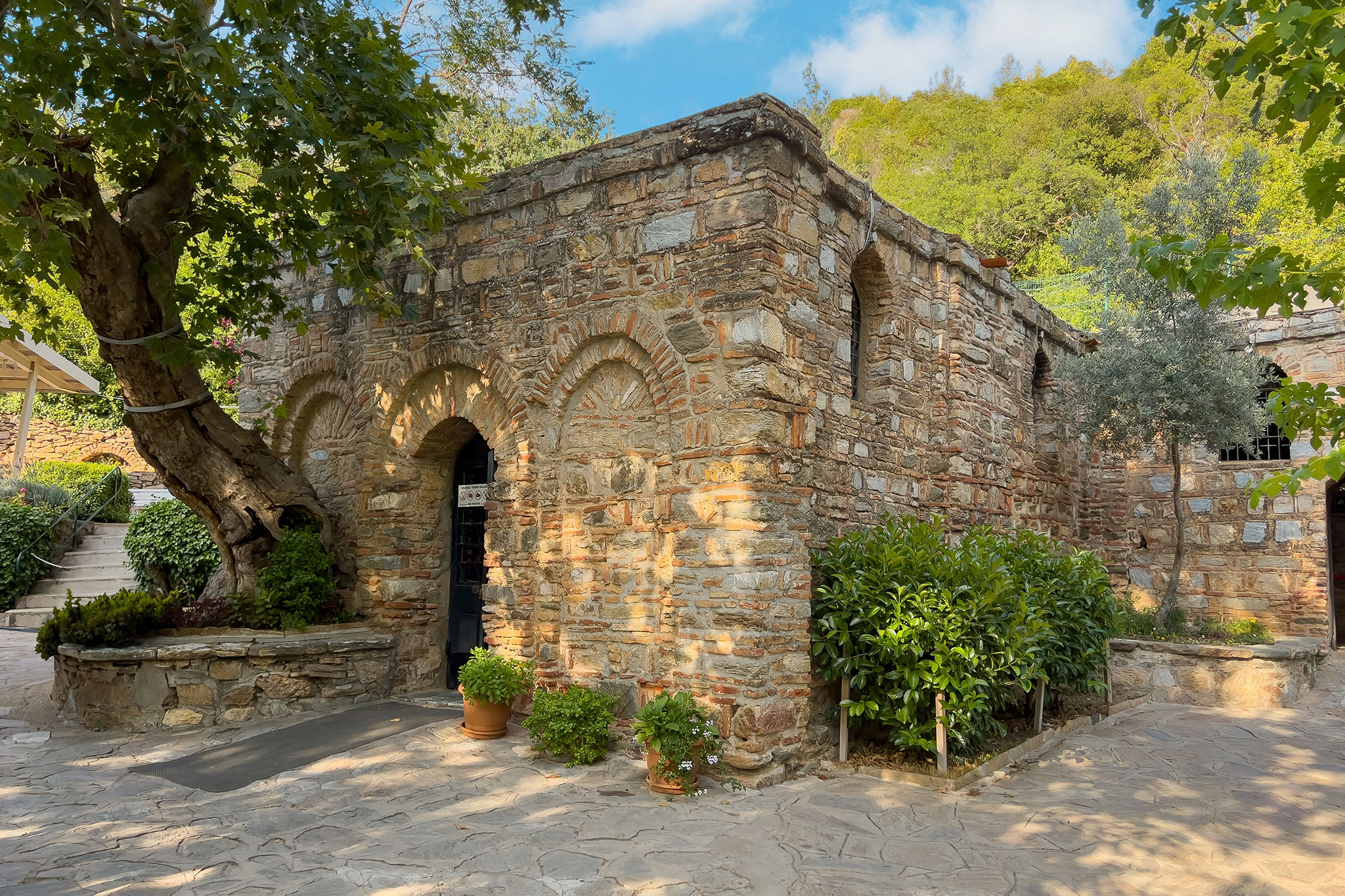 Historic stone house nestled among plants with tree branches overhanging the arched entrances.