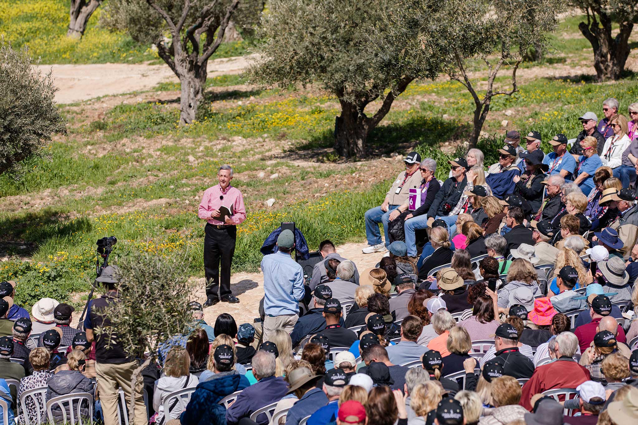 Dr. Robert Jeffress addresses a crowd of tourists in a field dotted with olive trees on a sunny day.