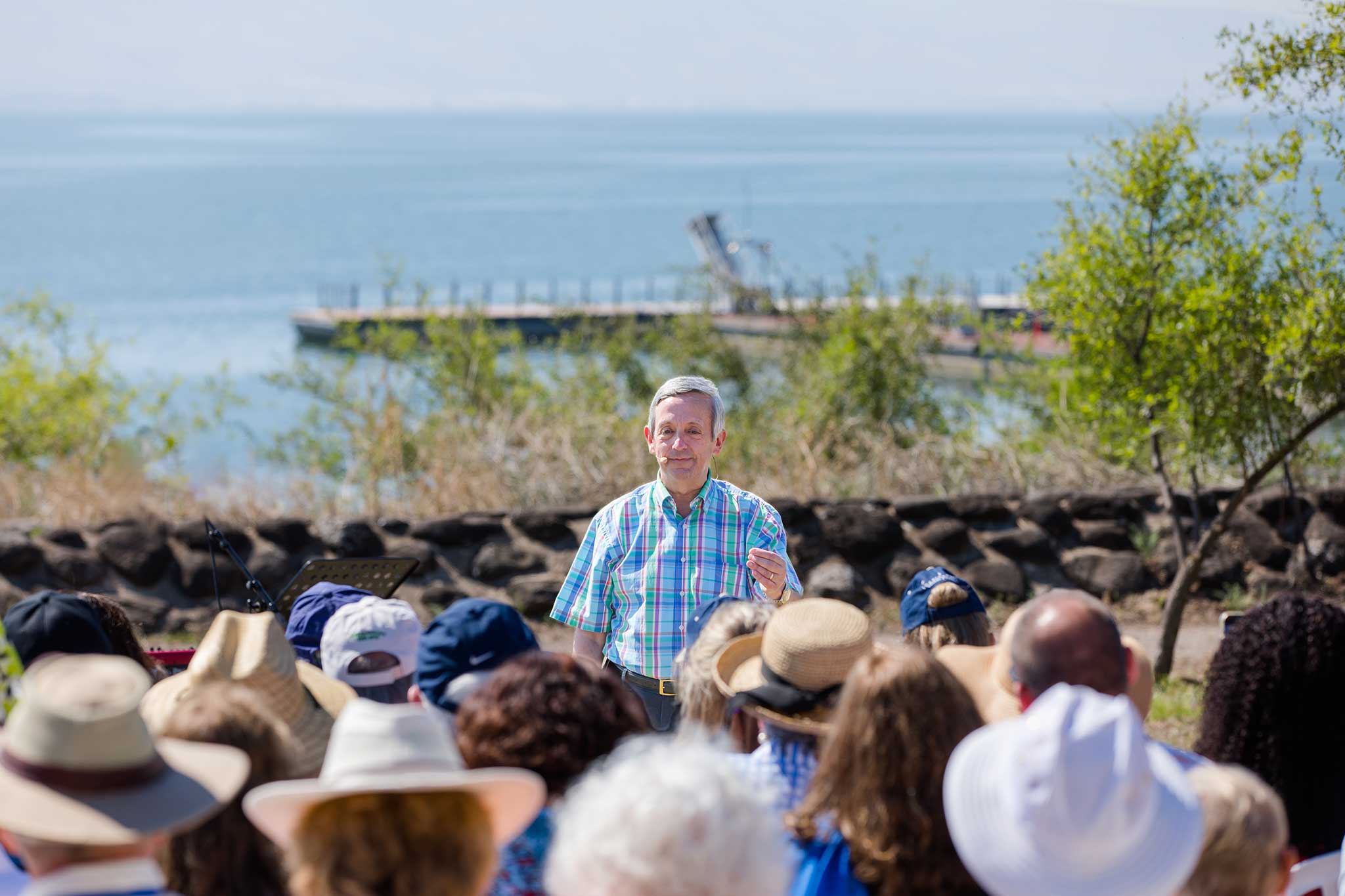 Dr Robert Jeffress lecturing to a group of tourists wearing hats, with a serene lake and a small dock in the background.