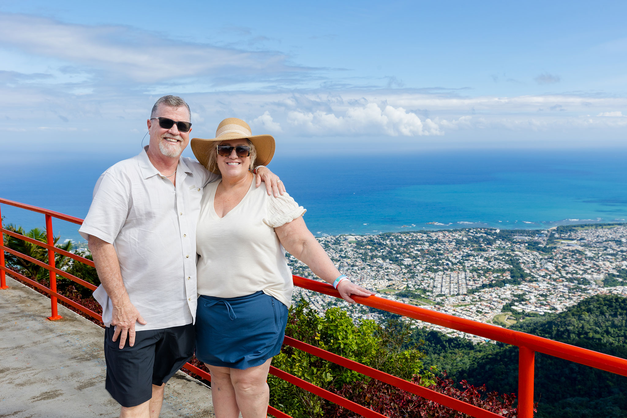 A man and woman in casual wear standing by a red railing overlooking a vast city and seascape.