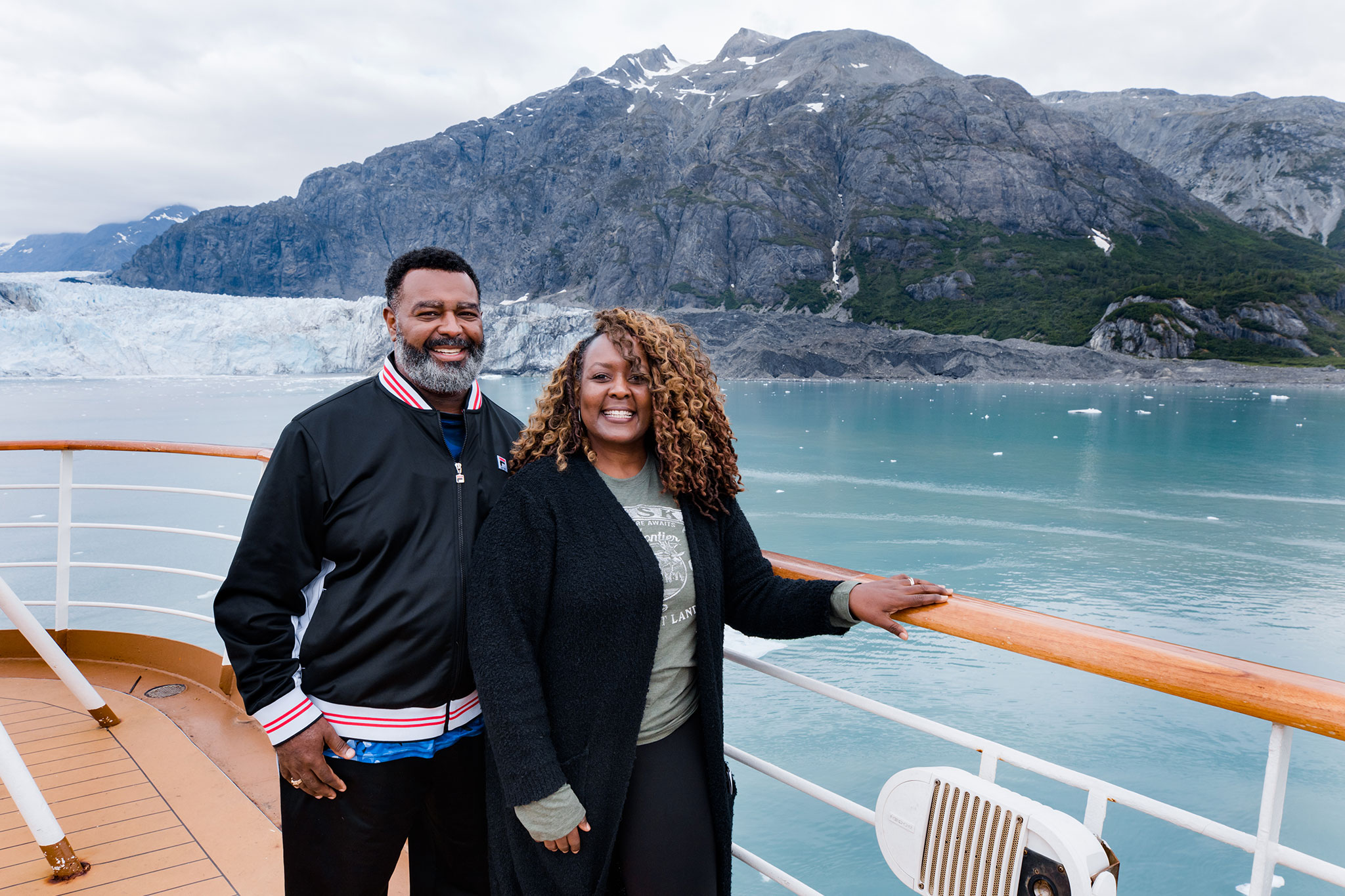 Middle-aged couple smiling on a cruise ship deck with a glacier and mountains in the background.