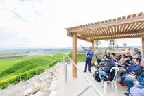 A man holding a book and gesturing toward a panoramic view of lush hills and fields, with tourists seated under a pergola listening intently.