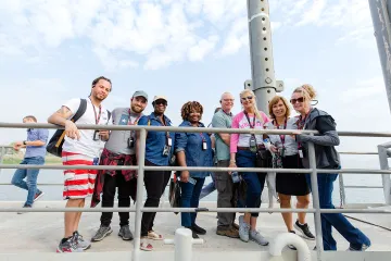 A cheerful gathering of travelers consisting of men and women of various ages and ethnicities on a bridge, with the sea visible beneath a cloudy sky in the background.